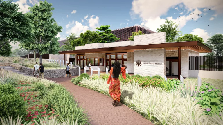 Exterior daylight rendering of the consular building and American Center entry at the U.S. Embassy in Lilongwe featuring prominent signage, landscaping, trees, a footpath with five visitors, and a retaining wall with adjacent steps