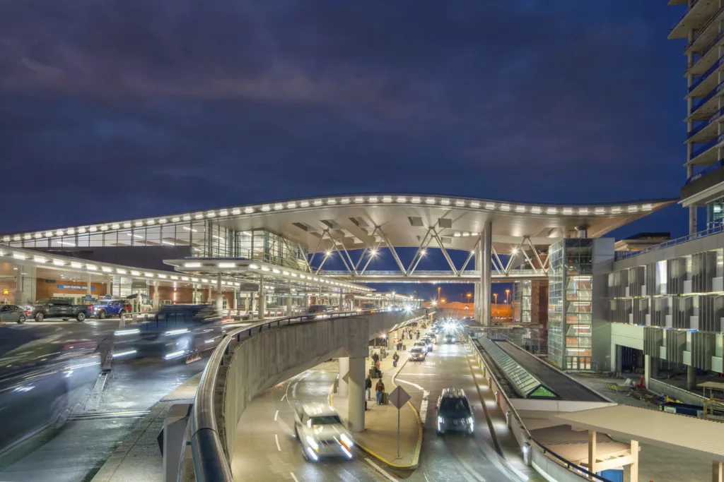 Exterior twilight image of the curvilinear airwave roof and pedestrian skybridge connected to Nashville International Airport's Terminal Lobby and Arrivals Facility, with arriving and departing vehicles dropping off and picking up travelers