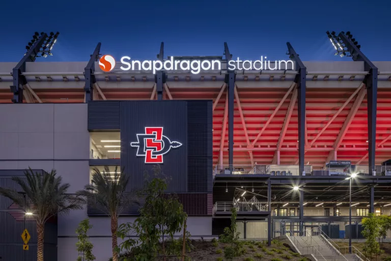 Exterior twilight view of the illuminated signage mounted to San Diego State University’s Snapdragon Stadium, with a staircase leading to the entrance and surrounding palms trees and landscaping