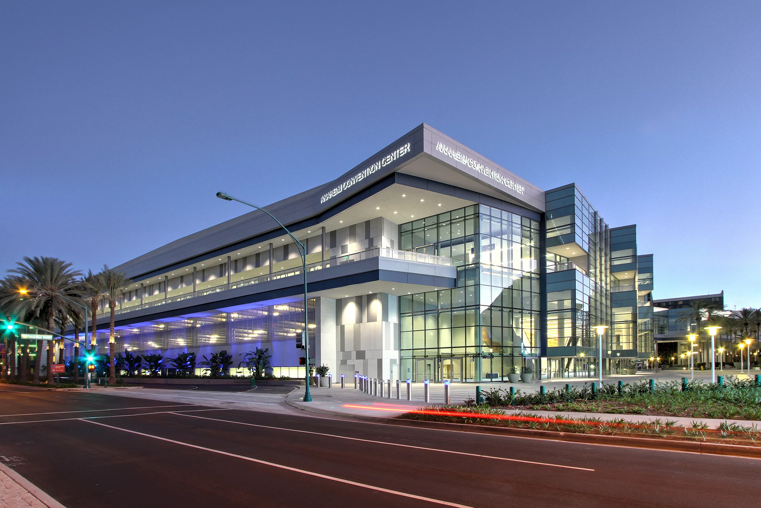 Exterior twilight view of the Anaheim Convention Center Expansion illuminated from within, featuring a large outdoor plaza with bollards, landscaping, and decorative street lamps