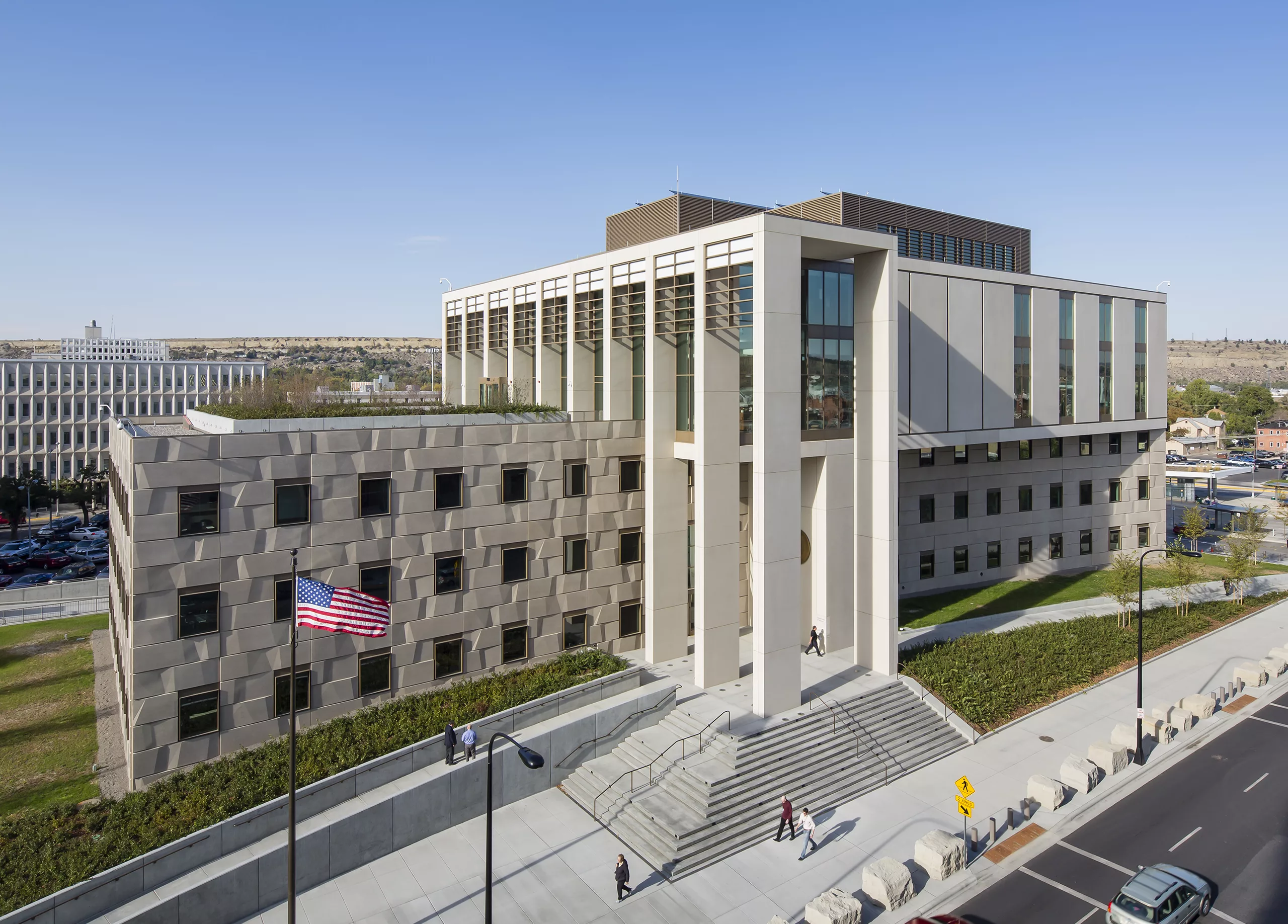Aerial daylight view of the six-story James F. Battin United States Courthouse featuring rooftop photovoltaic panels, steps leading to the entrance, sidewalk landscaping, and stone bollards adjacent to a two-lane road