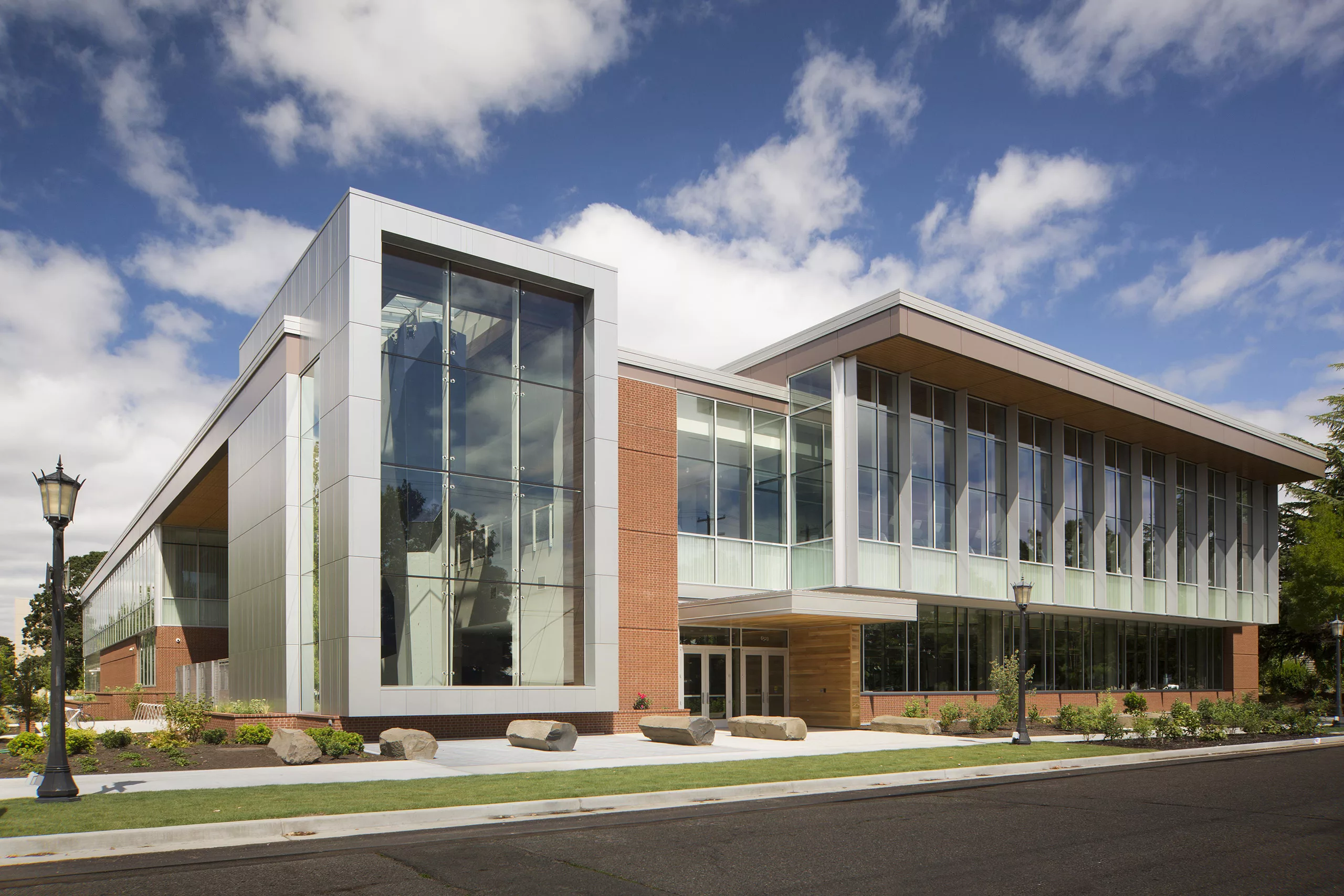 Exterior daylight view of the University of Portland's two-story Beauchamp Recreation and Wellness Center with a climbing wall and exercise equipment inside and boulder-shaped benches, landscaping, and a parking lot outside