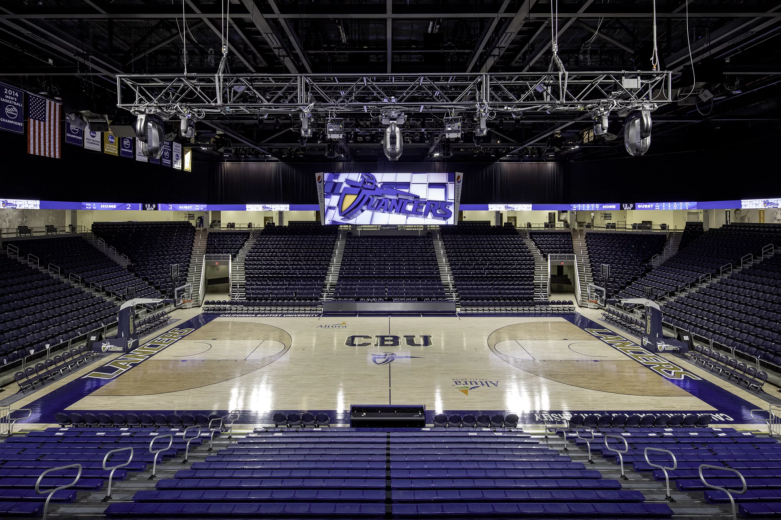 Interior view of the California Baptist University Arena's basketball court featuring overhead rigging, a video board, and bleacher seating