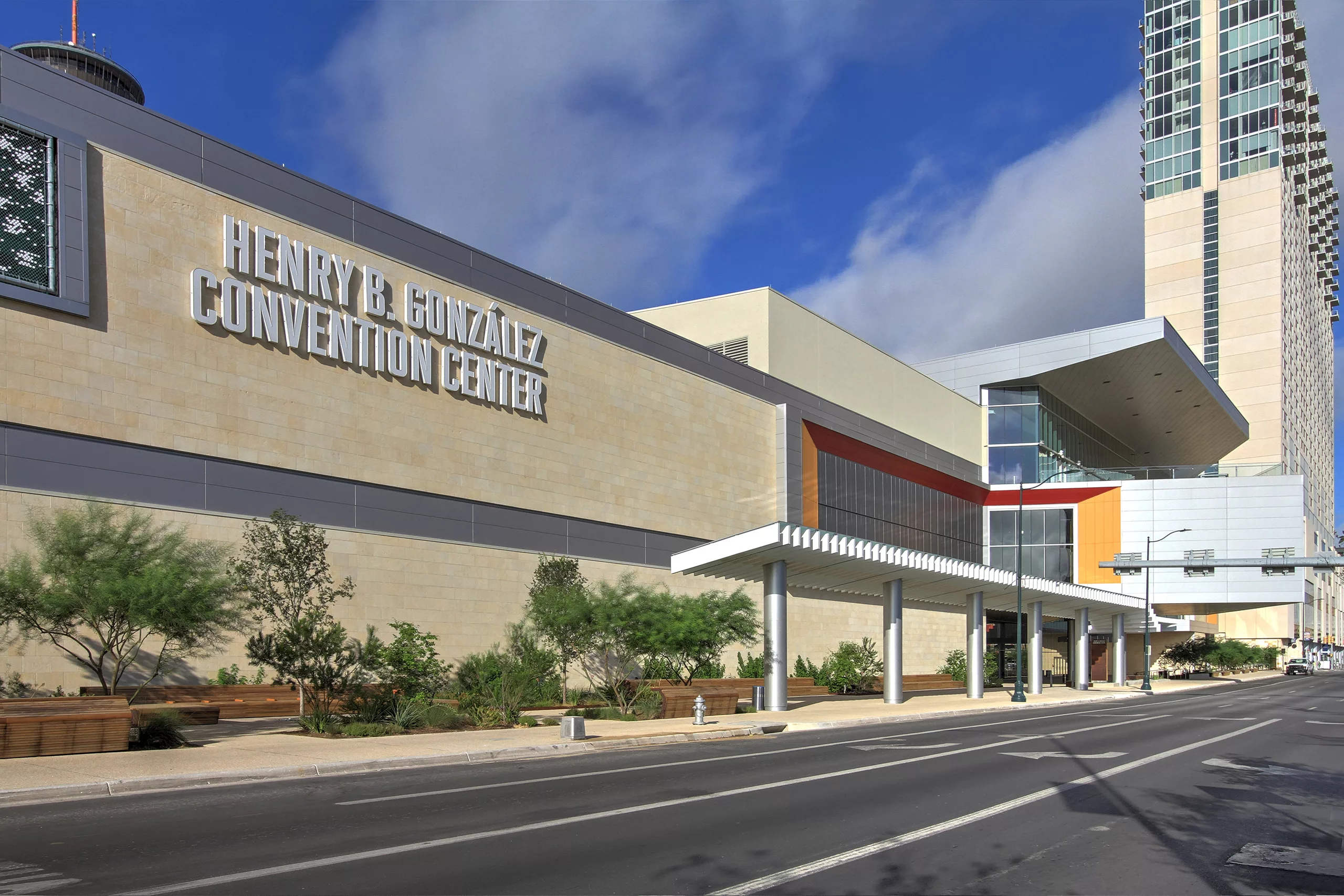 Exterior daylight view of the Henry B. González Convention Center Expansion with a roadway and pedestrian canopy in front and a high-rise building beyond