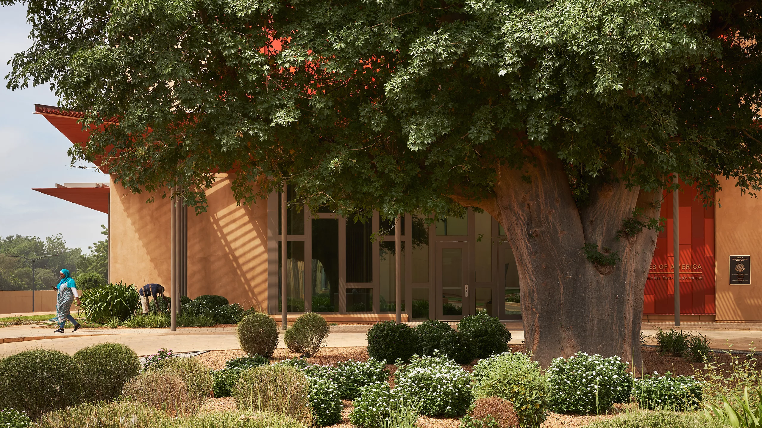 Exterior daylight view of the landscaped entrance to the U.S. embassy in Niamey with a large tree in front and one person leaving the building