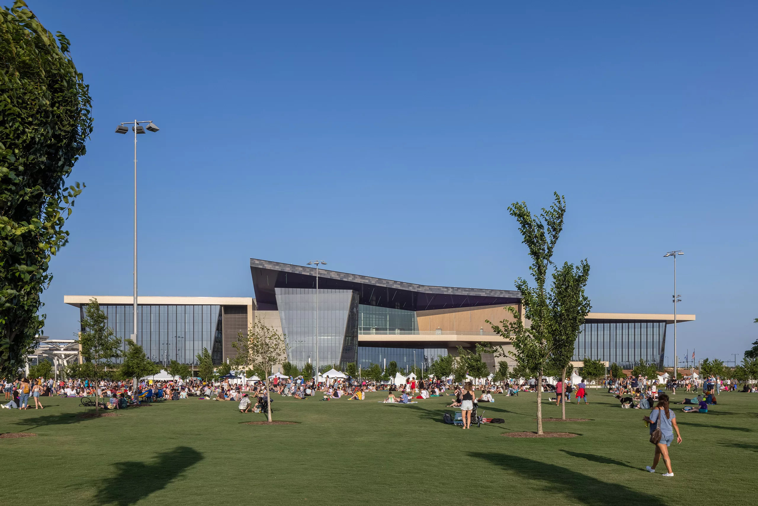 Exterior daylight view of the Oklahoma City Convention Center with an expansive front lawn dotted with trees, special-event tents, and people relaxing and recreating