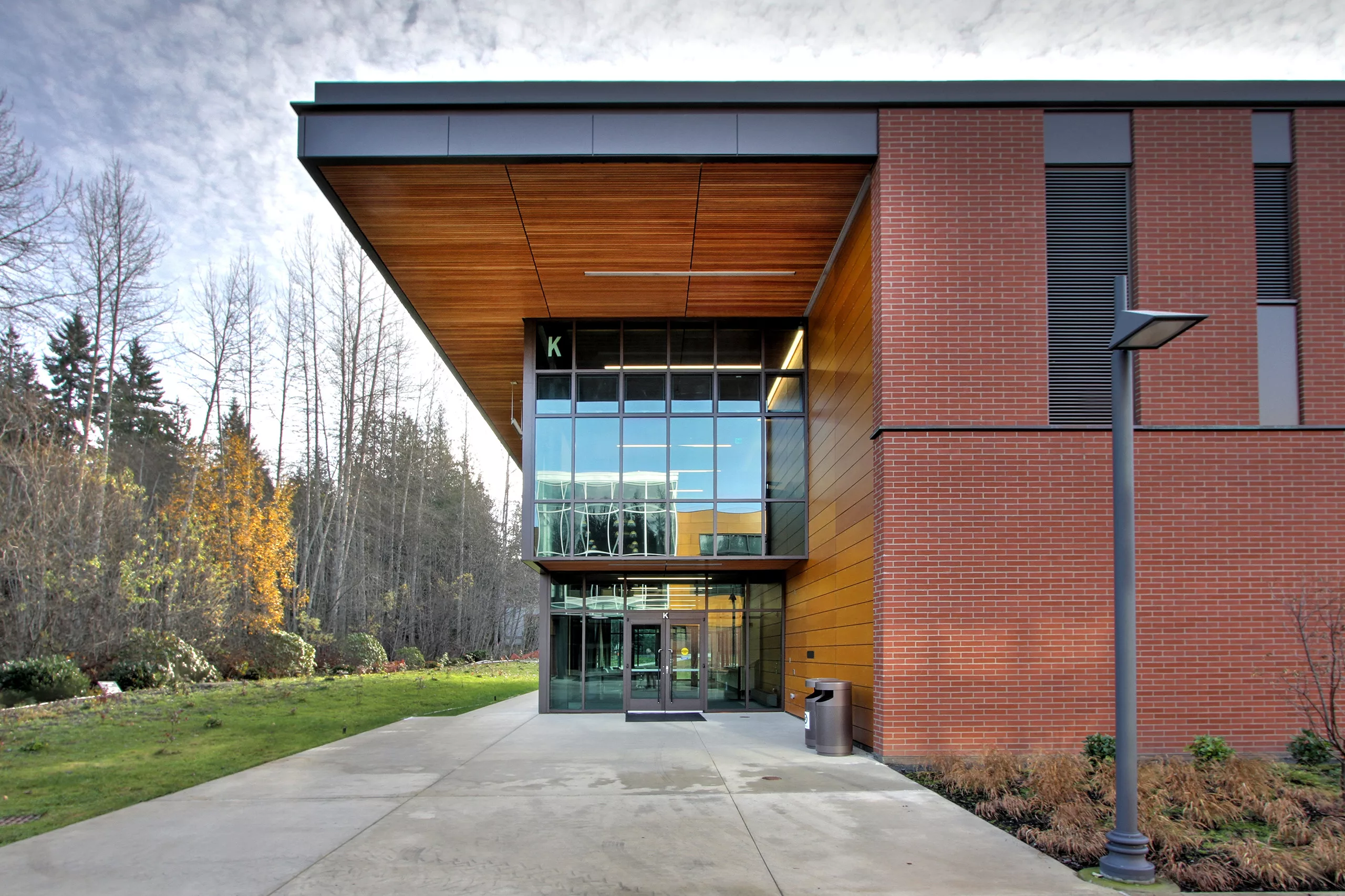 Exterior daylight view of the entrance to Peninsula College's Allied Health and Early Childhood Education Building with a pedestrian walkway, lamp post, and surrounding natural landscape