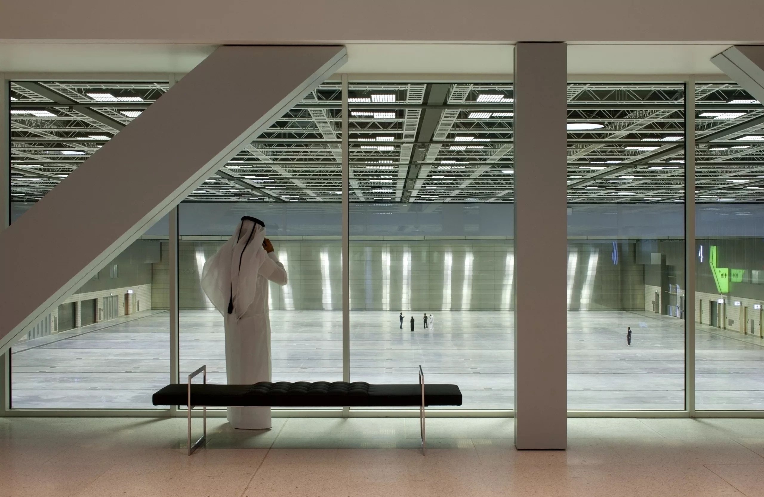 A man wearing a traditional white thobe stands in front of a glass wall with views of the expansive exhibit hall with overhead lights and five people standing on the floor below