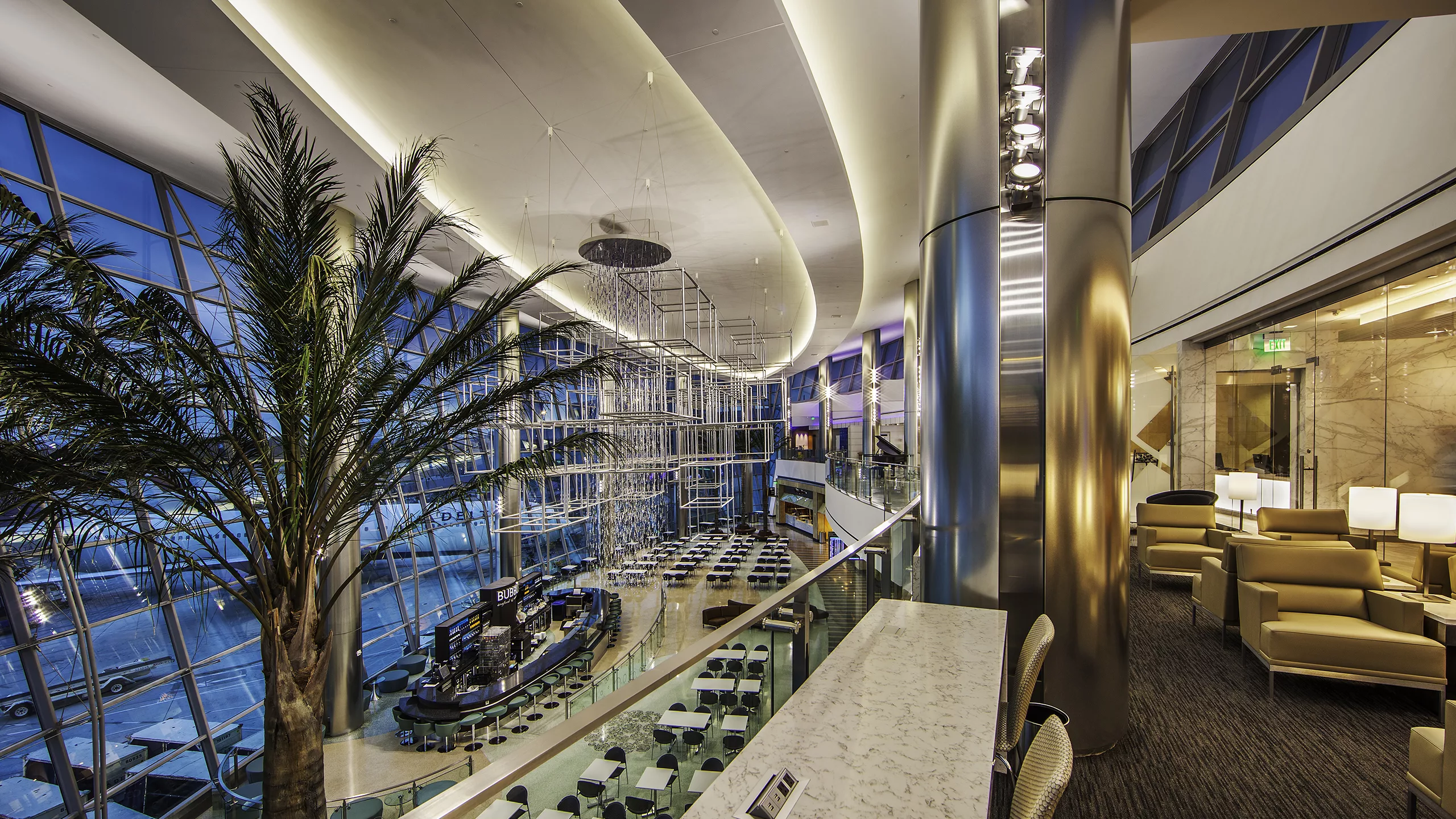 Interior evening image of the downstairs dining hall and an upstairs lounge with an indoor palm tree and ceiling-mounted, cube-shaped public art at San Diego International Airport Terminal 2 West