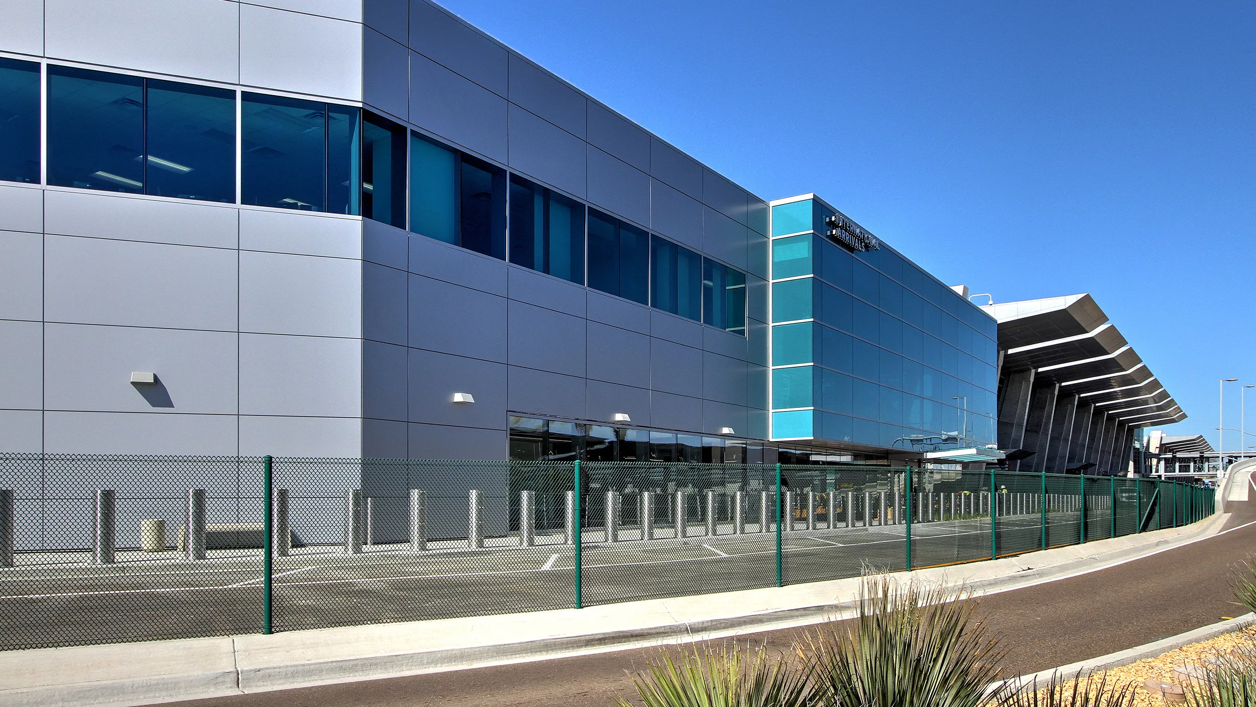 Exterior daylight view looking east toward San Diego International Airport's multi-story, glass-and-steel Federal Inspection Station fronted by steel safety bollards and minimal landscaping