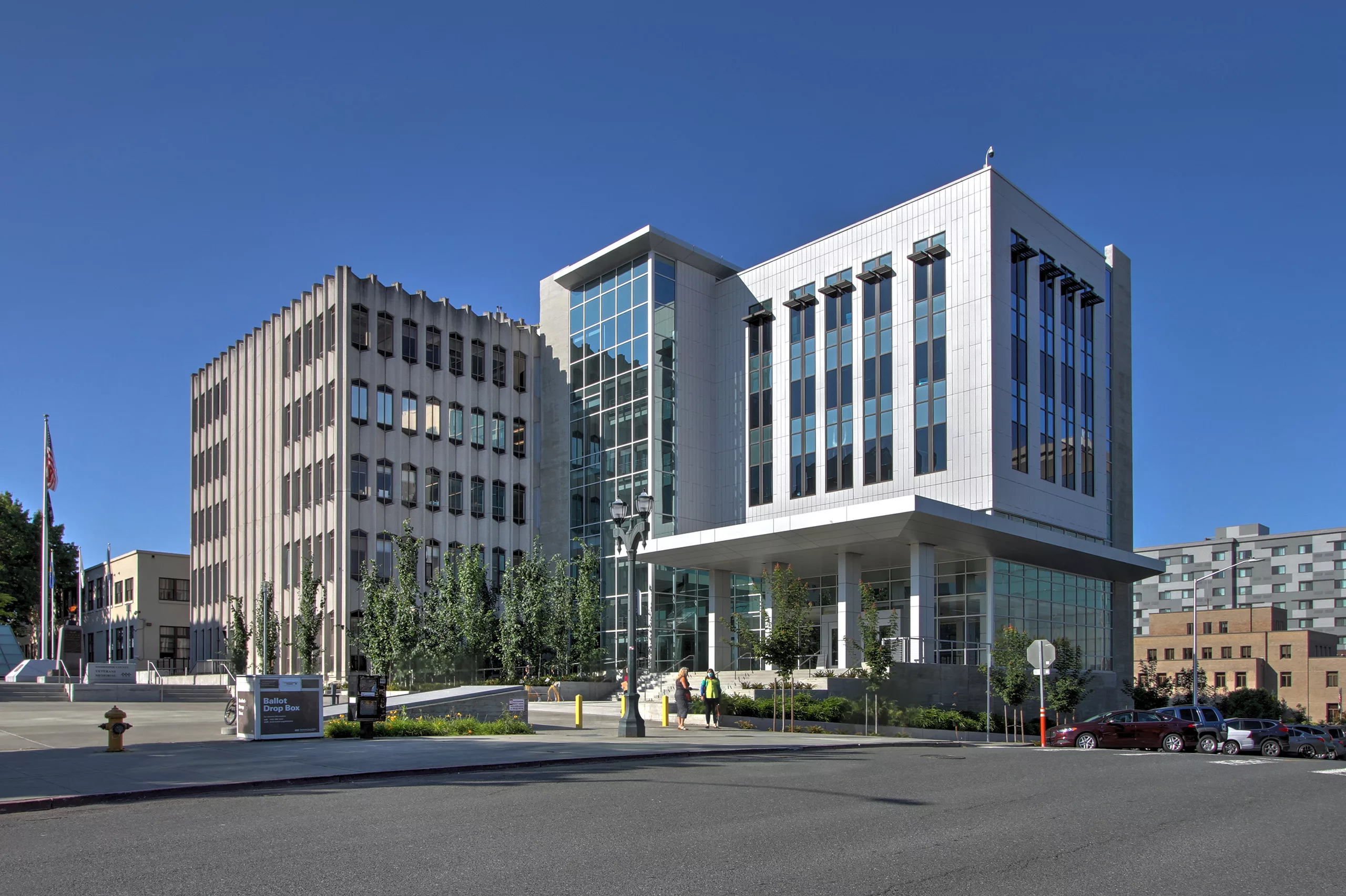 Exterior daylight view of Snohomish County Justice Center and its five-story addition with two people gathered outside the steps leading to the entrance and a large ballot drop box, newspaper stand, and landscaped sidewalk