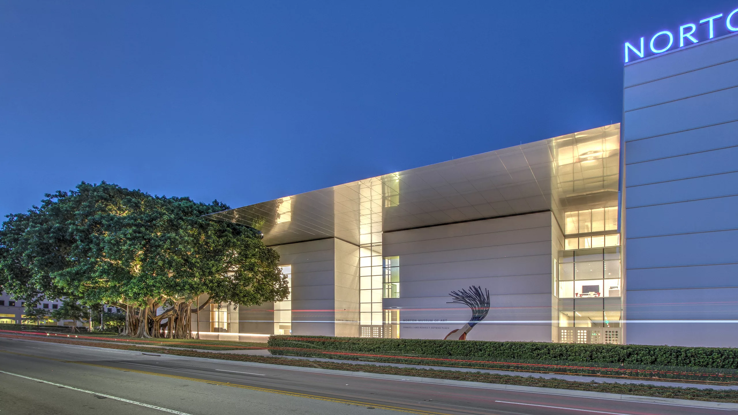 Exterior twilight view of the illuminated steel-and-glass entrance to the Norton Museum of Art Expansion, with bonsai tree and outdoor public art